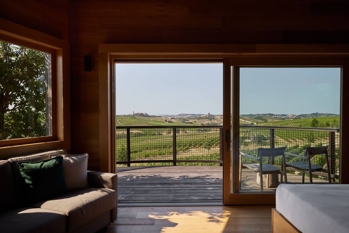 Une pièce avec un plafond en bois, un canapé agrémenté d’un coussin vert, un lit et un balcon offrant une vue sur des vignobles et une ville lointaine.