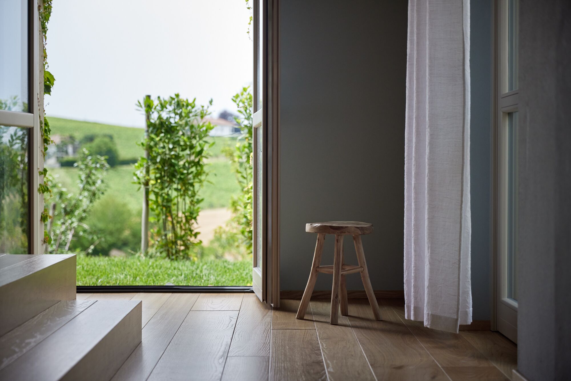 A room with a wooden floor, a wooden stool, and a view of a green landscape through a window.