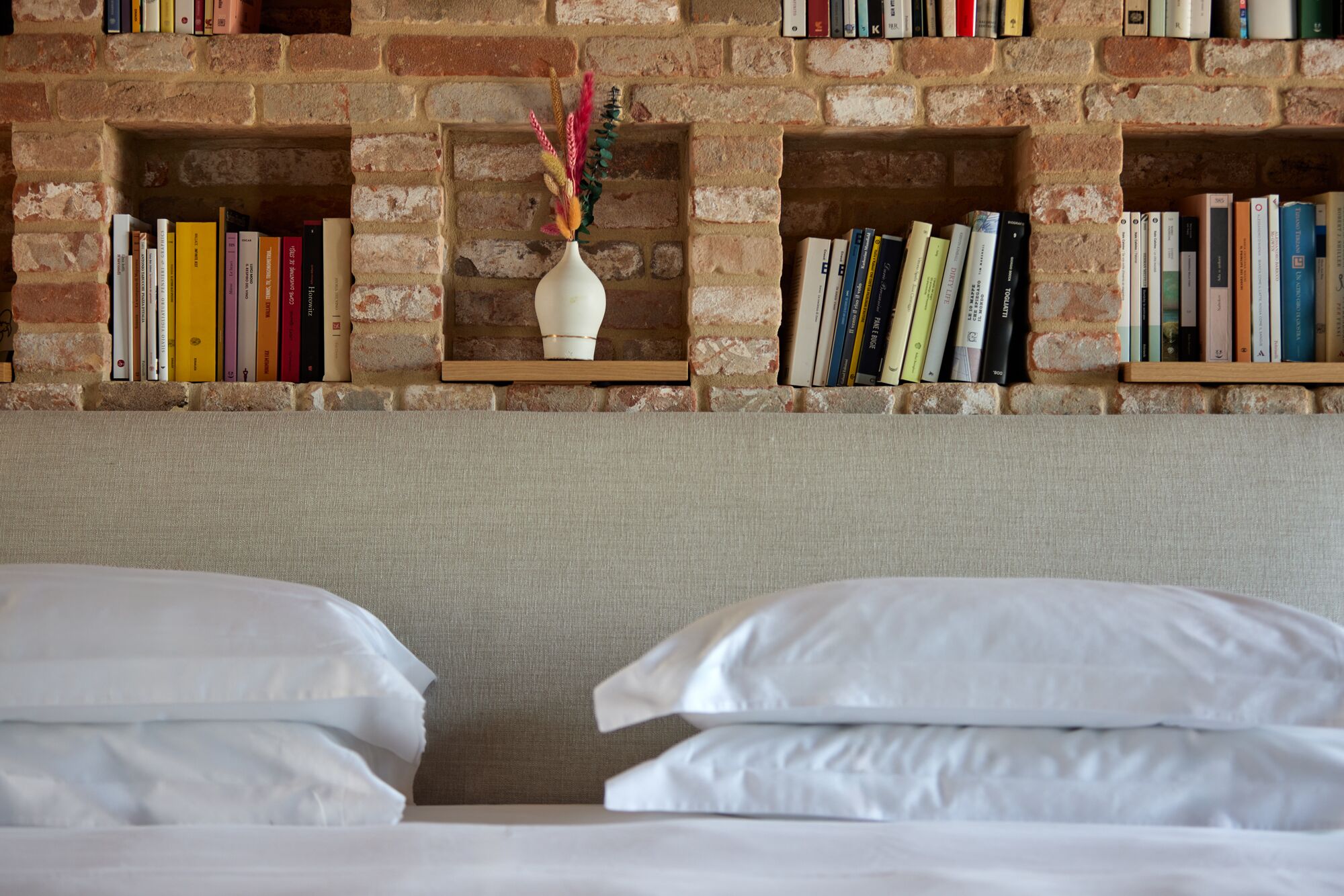 Habitación de hotel con pared de ladrillo, dos camas con ropa blanca y estanterías con libros y un jarrón decorativo.