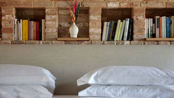 Habitación de hotel con pared de ladrillo, dos camas con ropa blanca y estanterías con libros y un jarrón decorativo.