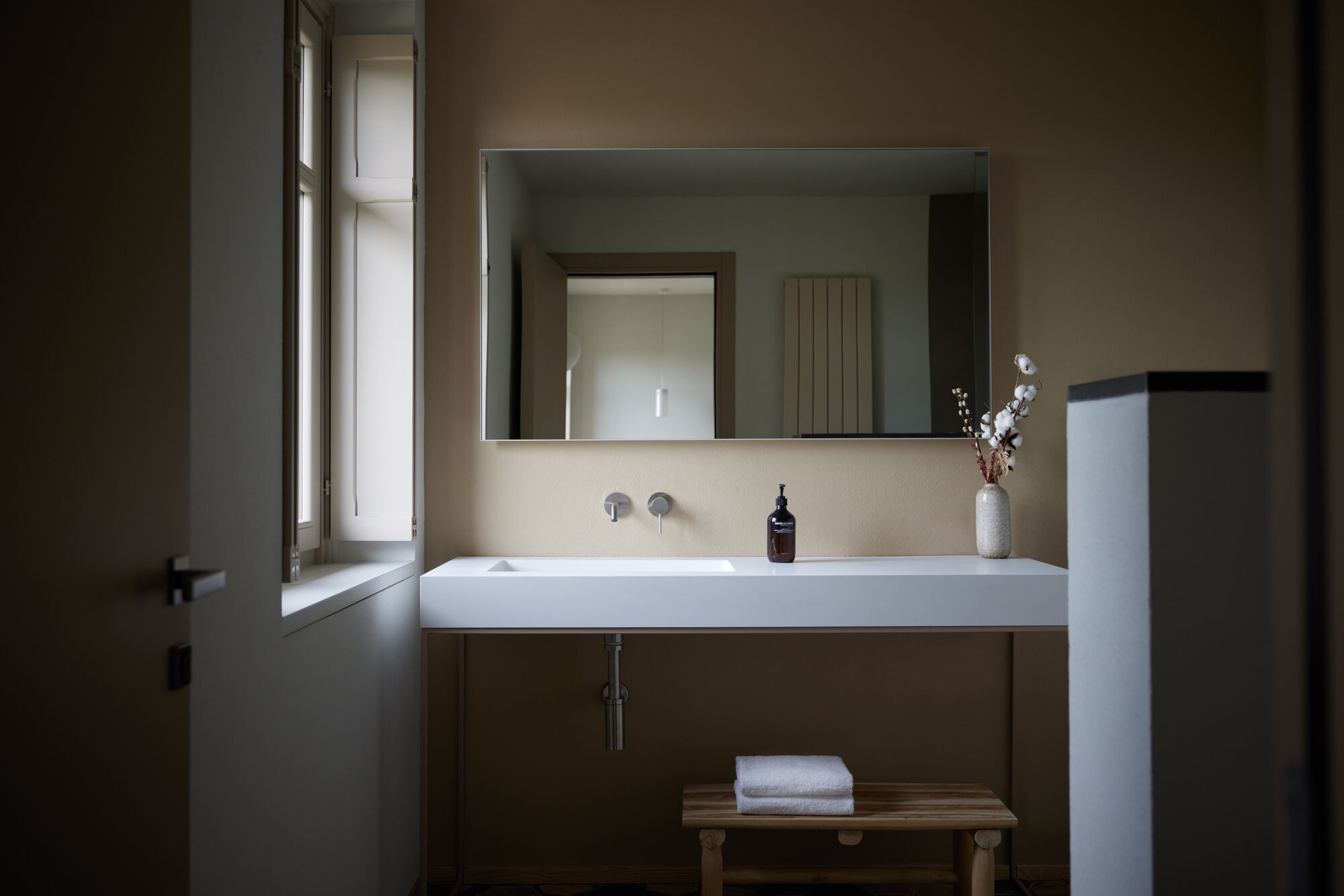 Un baño con un lavamanos blanco, un espejo, una botella de jabón y un pequeño banco de madera con una toalla.