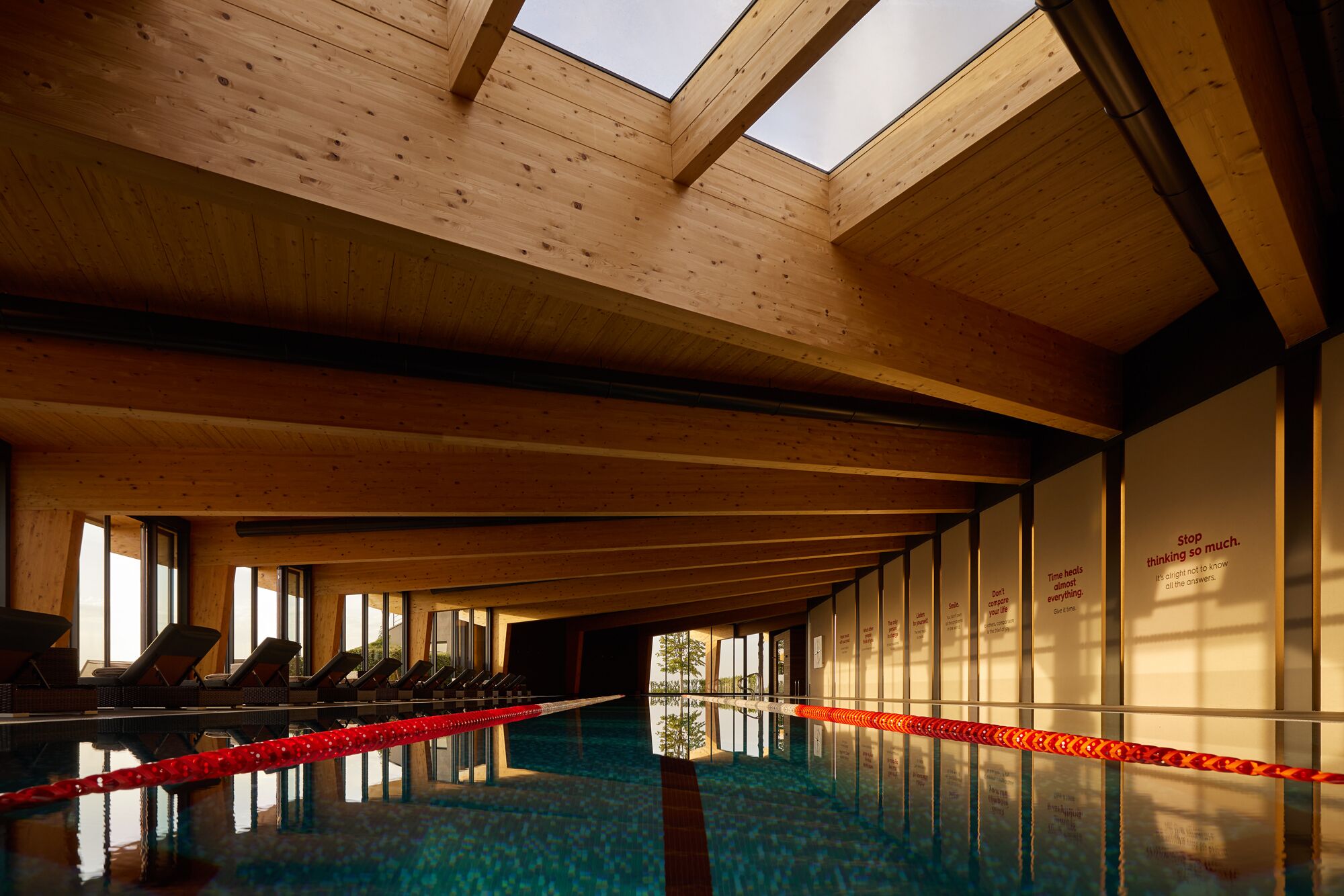 A swimming pool with wooden beams and skylights, surrounded by lounge chairs and motivational quotes on the walls.