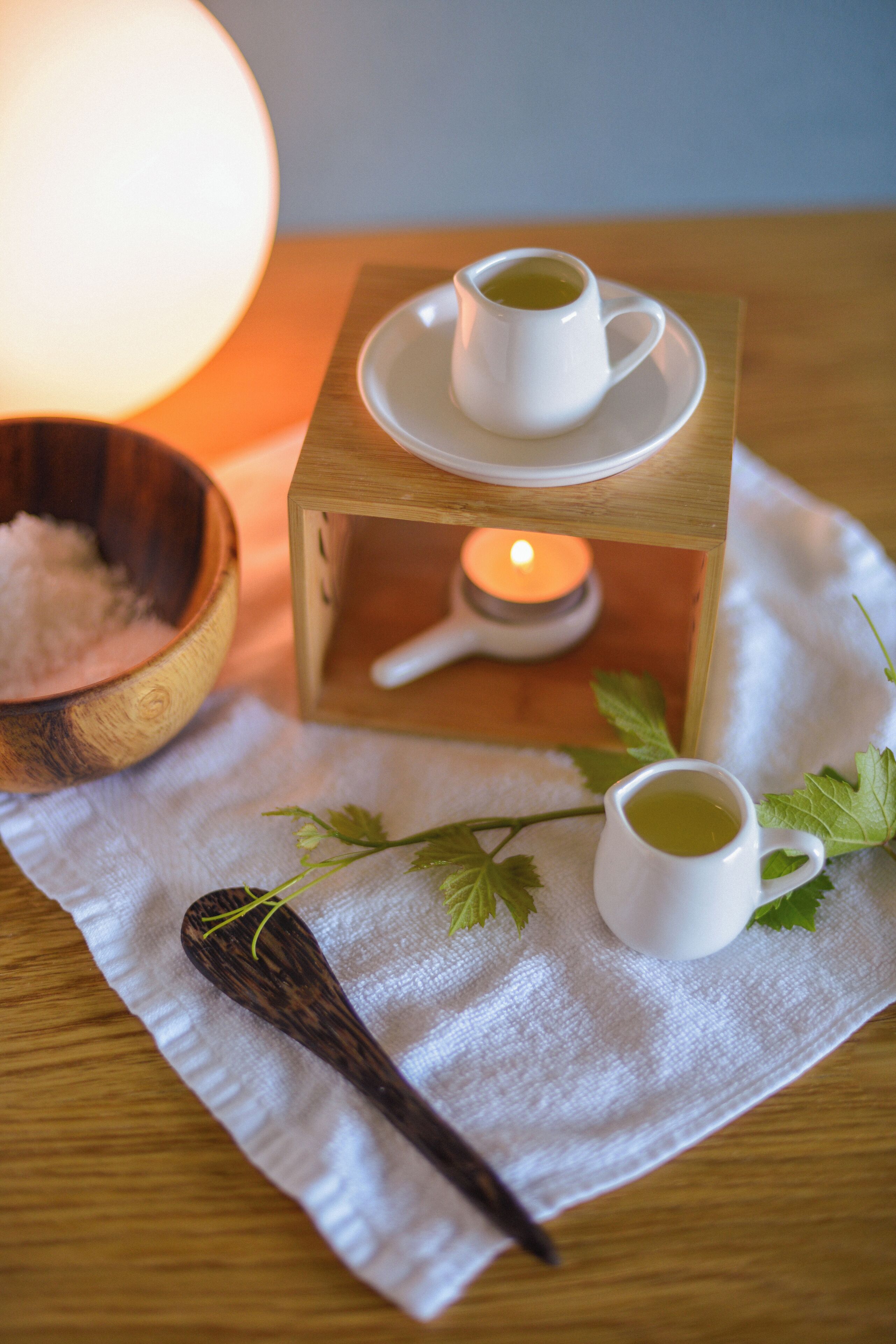 A wooden table with two white cups of tea, a wooden bowl, a lit candle, and a spoon on a white cloth.