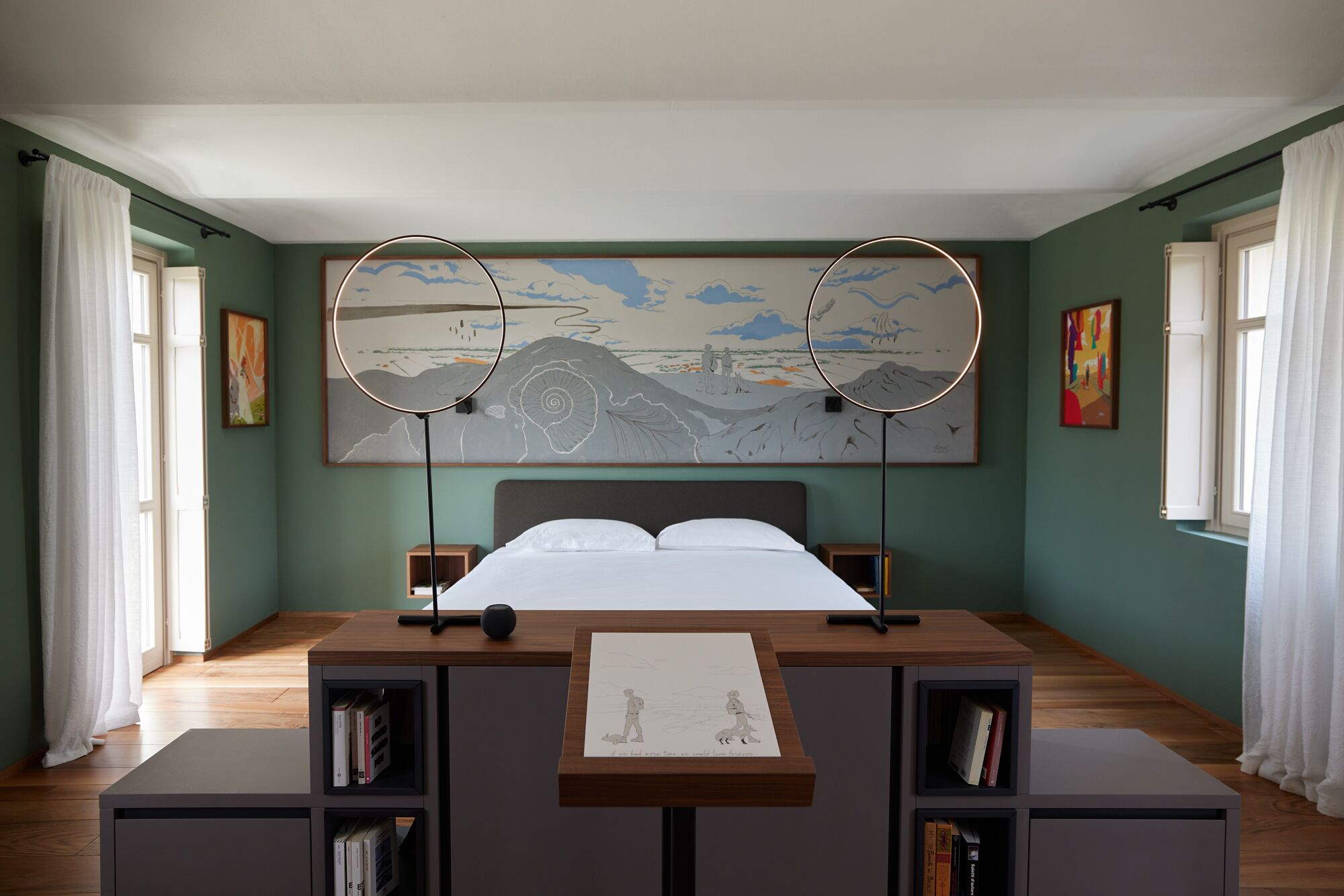 A bedroom with a bed, two bedside tables, a large artwork above the bed, and a desk with a chair in the center.