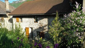 A rustic stone building with a sloped roof, surrounded by lush greenery and purple flowers. - Artist'au Chat" family gîte with sauna (saint paul)