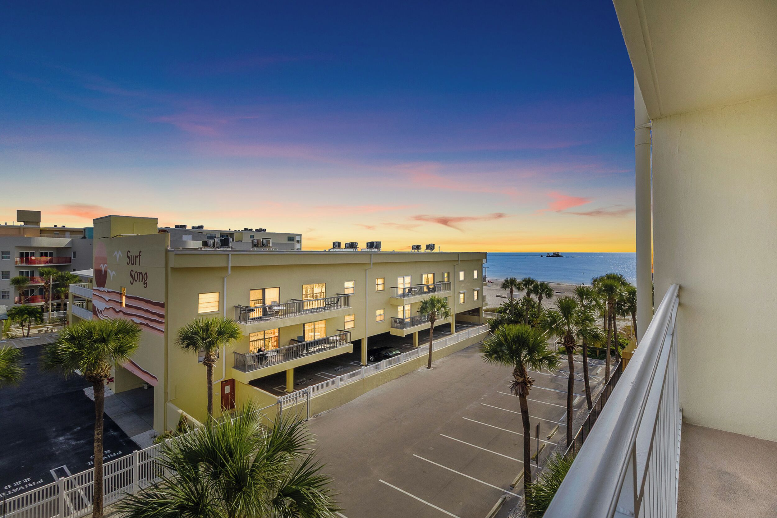 A hotel with a parking lot, multiple floors, and a view of the beach and ocean at sunset.