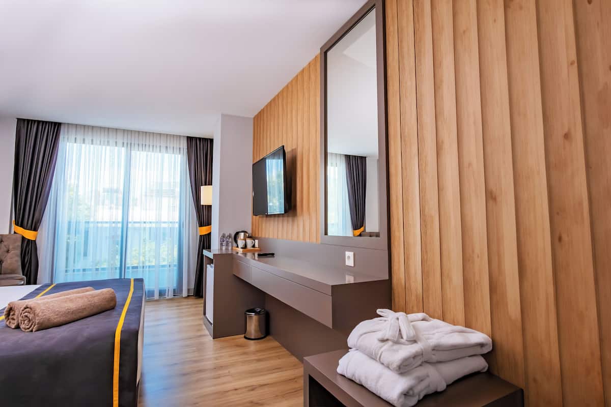 A modern hotel room with a bed, a TV, a wooden panel wall, a mirror, and a window with curtains.