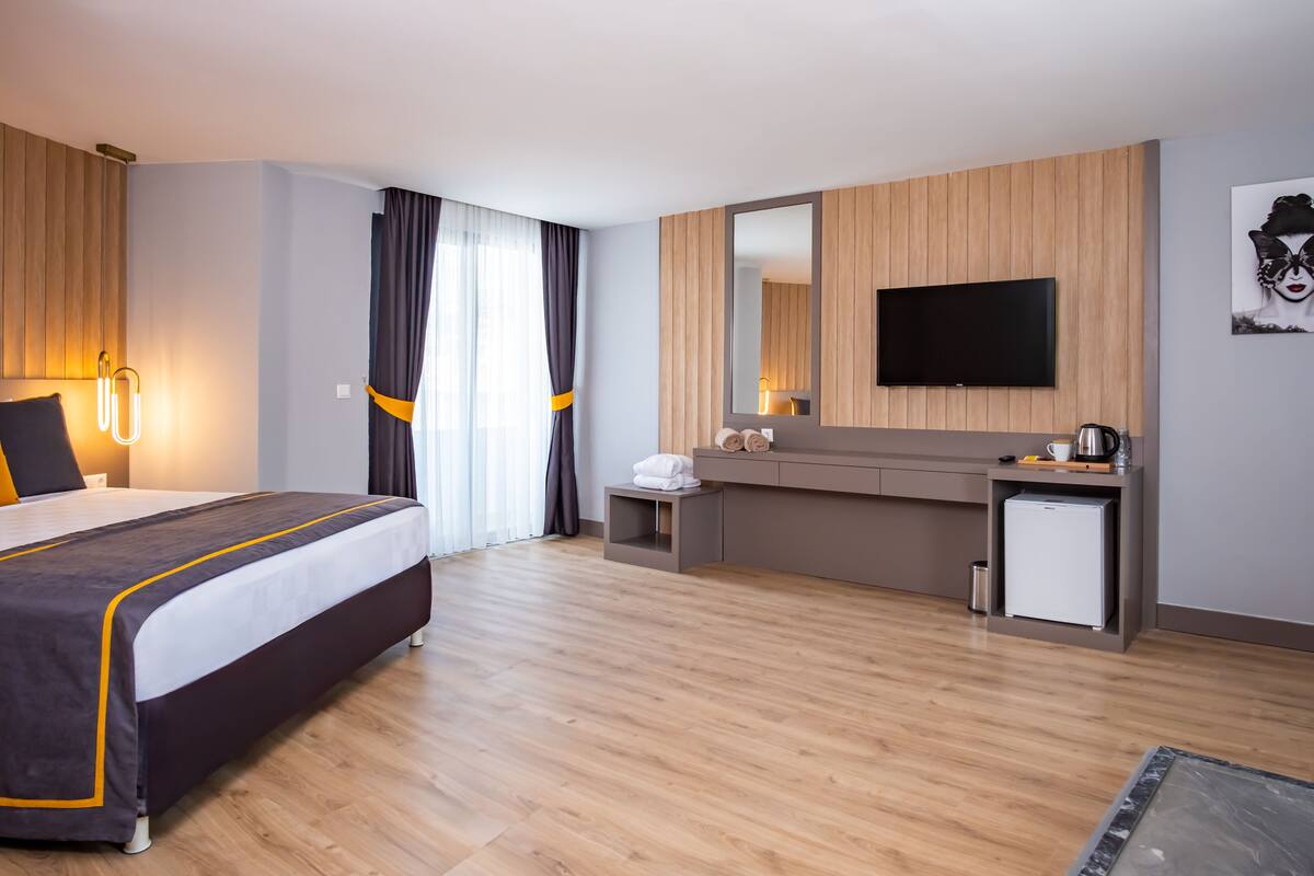A modern hotel room with a large bed, a small desk, a flat-screen TV, and a compact kitchenette.