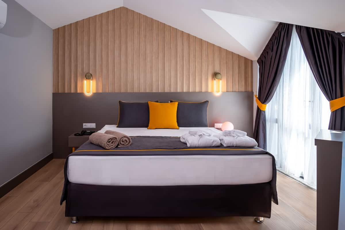 A modern hotel room with a large bed, wooden panel wall, two bedside lamps, a nightstand, and a window with curtains.