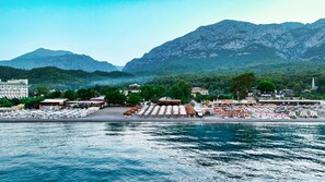Un complejo turístico costero con asientos frente a la playa, un puerto deportivo y montañas al fondo.