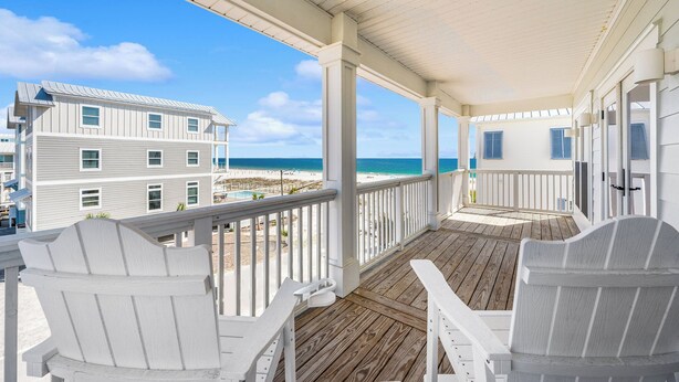 Un porche cubierto con sillas Adirondack blancas, una terraza de madera y vista a la playa y el océano.