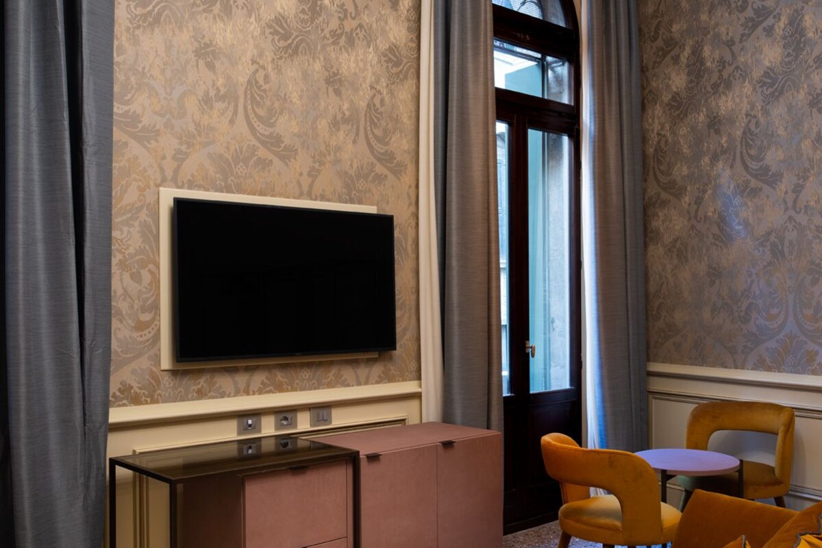 Habitación de hotel con una televisión de pantalla plana grande, un sofá, una mesita y una ventana con cortinas.