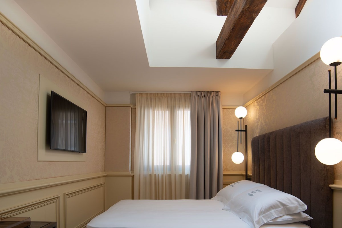 Hotelkamer met een bed, een flatscreen televisie aan de muur, een raam met gordijnen en een nachtkastje met een lamp.