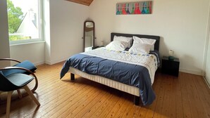 Quarto com teto de madeira, cama com colcha azul e branca, mesa de cabeceira com luminária, uma cadeira e janela com vista para árvores.