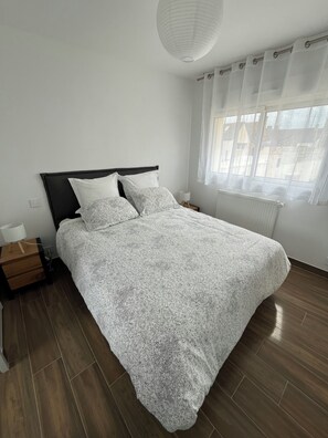 A bedroom with a bed, a nightstand, a lamp, a window with curtains, and a radiator.