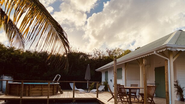 A wooden deck with lounge chairs and a pool, a small building with a porch, and a palm tree. - VILLA KAZ’ARY (Sainte-Anne)
