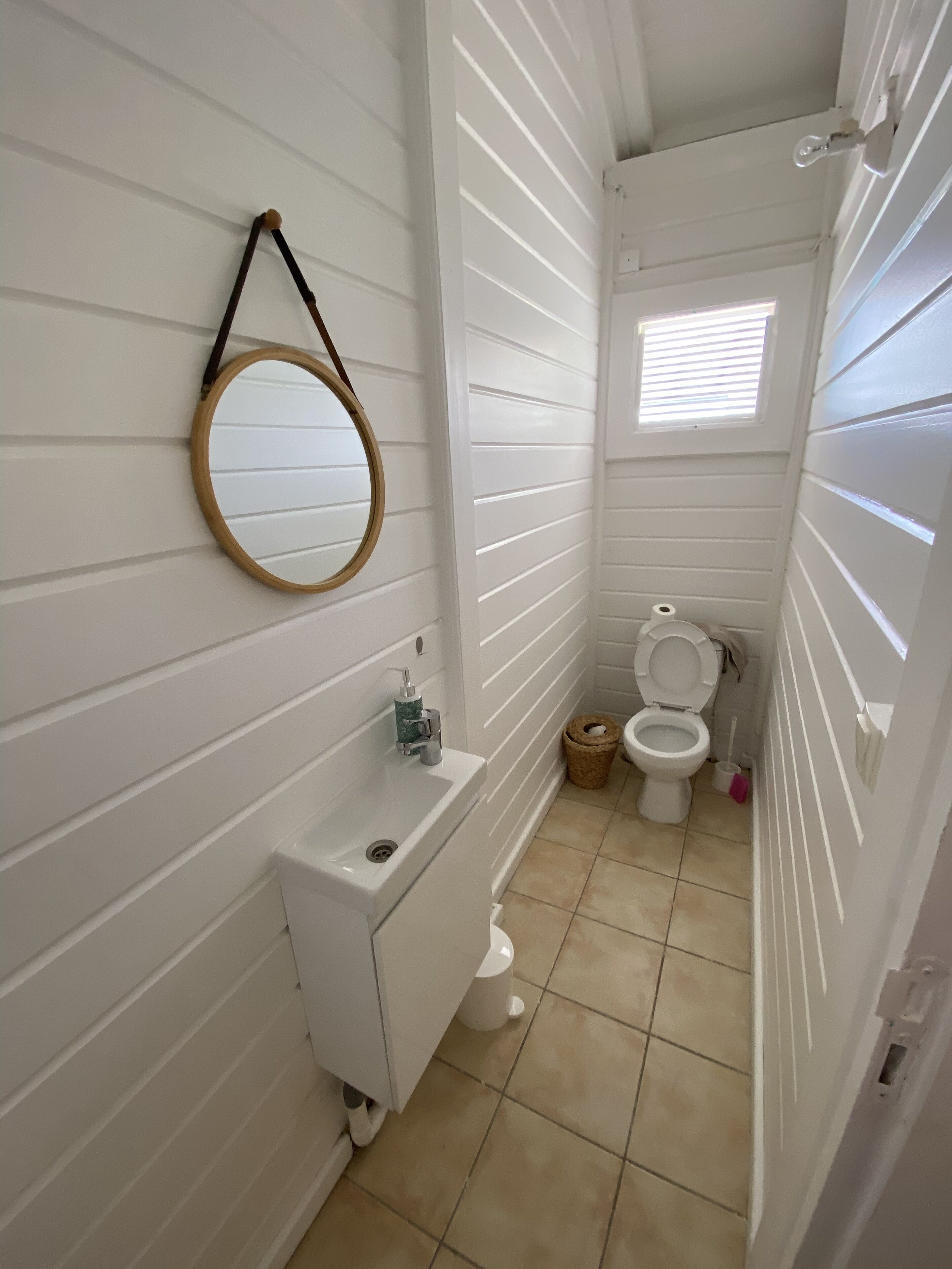 A small, narrow bathroom with white walls and a white sink. A round mirror hangs on the wall, and there is a window above the toilet.