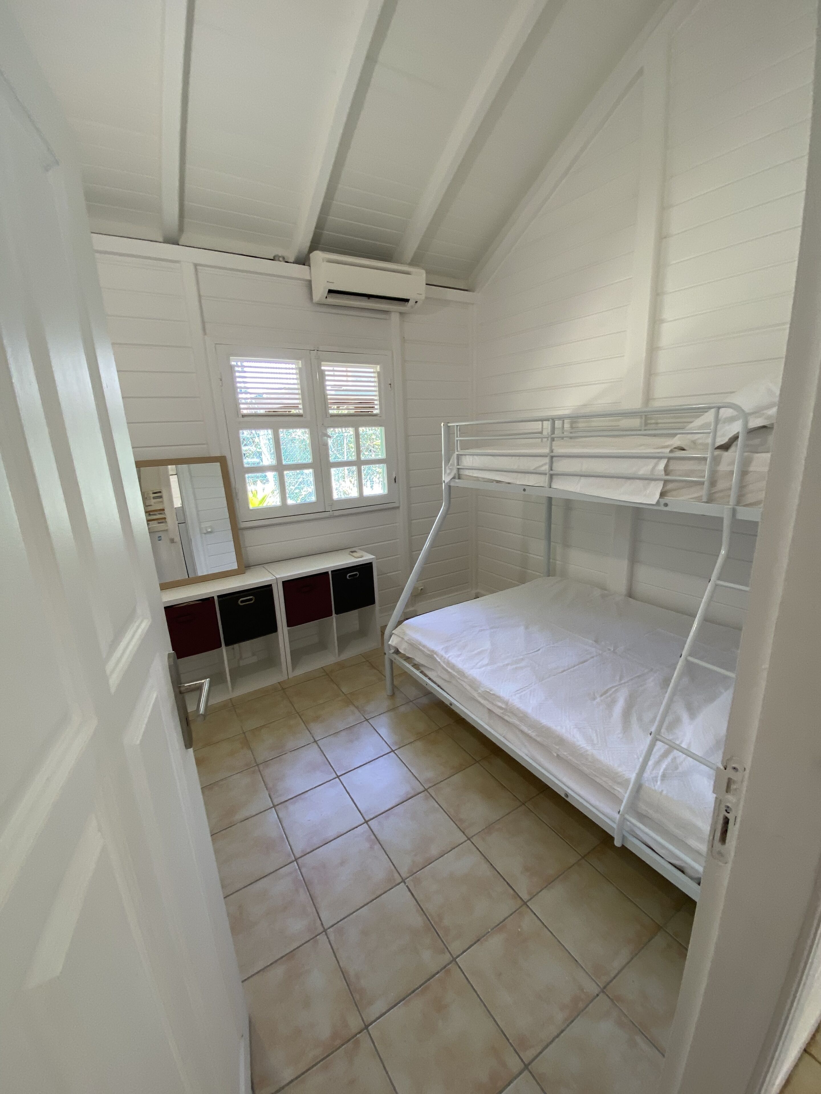 A dormitory room with bunk beds, a desk with drawers, a window with blinds, and an air conditioning unit.