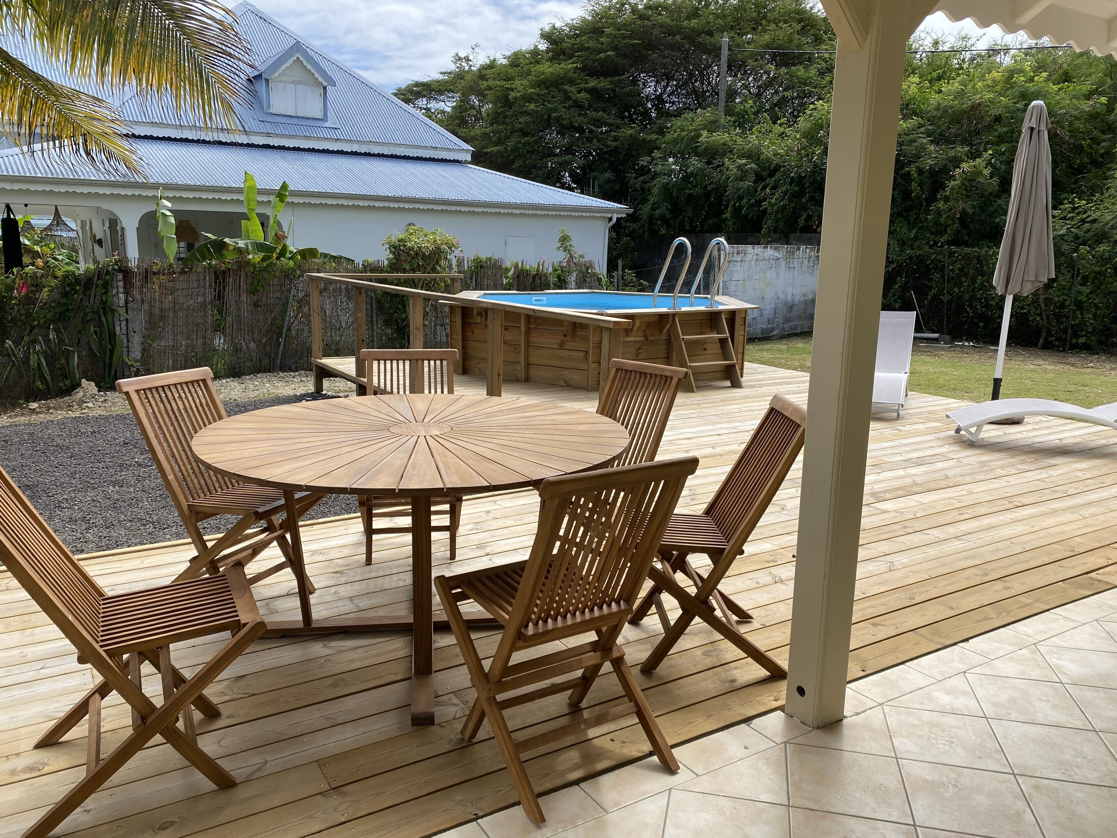 Een houten terras met een ronde tafel en stoelen, een zwembad en een overdekt gedeelte met een stoel en parasol.