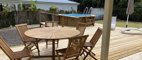 Een houten terras met een ronde tafel en stoelen, een zwembad en een overdekt gedeelte met een stoel en parasol.