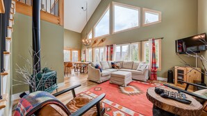 A spacious living room with a large sectional sofa, a wooden coffee table, and a red patterned rug. There are multiple windows providing natural light, and a television mounted on the wall. - 5BR Retreat w/ Hot Tub - Sleeps 12 in Stratton (Stratton)