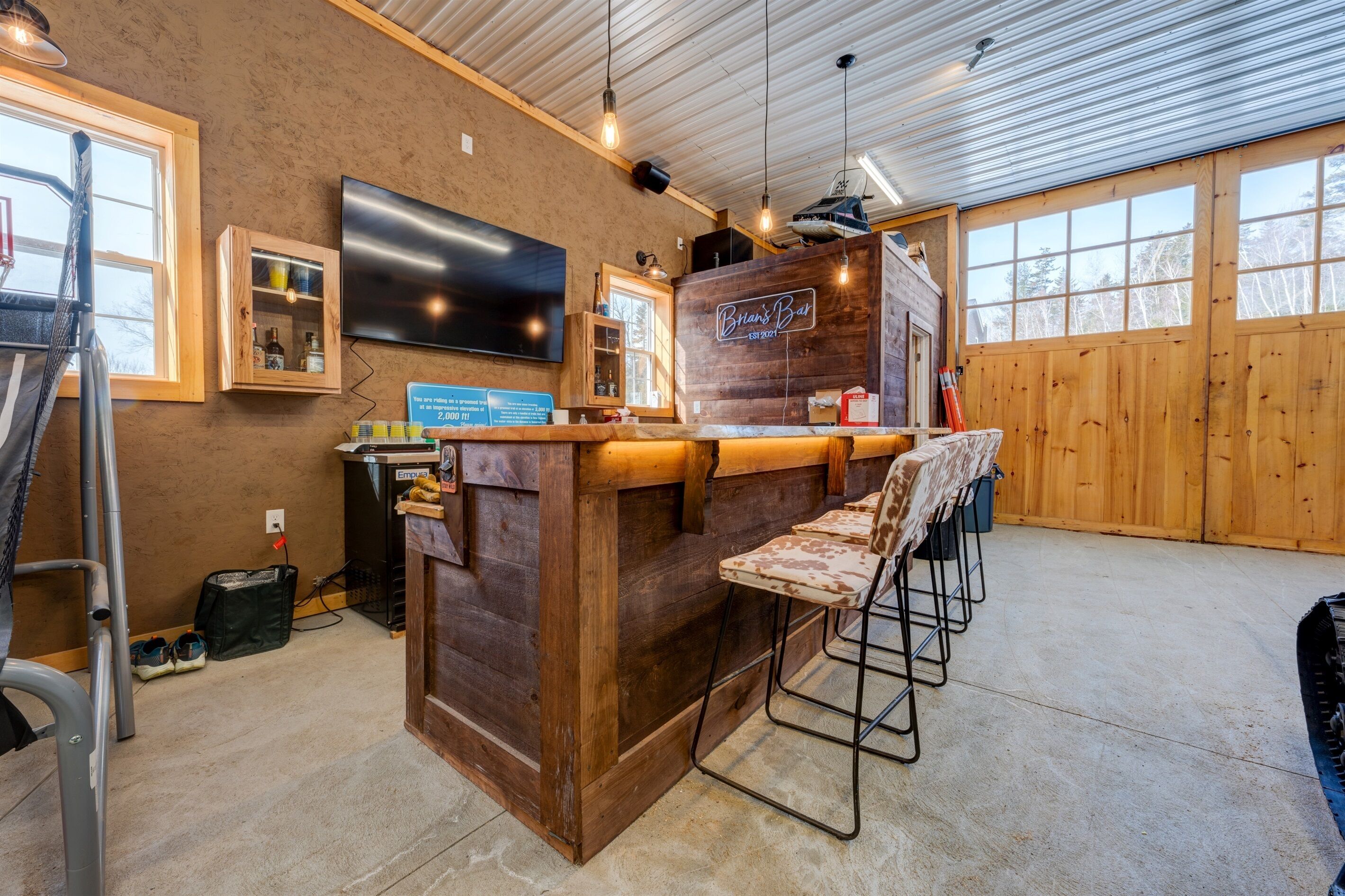 Una zona de bar rústica con paredes de madera y una gran pantalla de televisión. Hay taburetes de bar y una barra con un letrero que dice "Bilt's Bar".