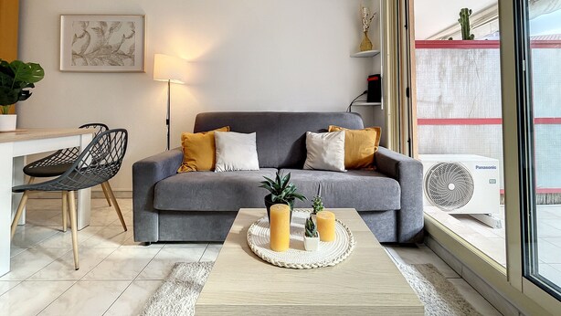 Un salon moderne avec un canapé gris, une table basse décorée de bougies et de plantes, un fauteuil noir, une lampe et une grande fenêtre offrant une belle vue.