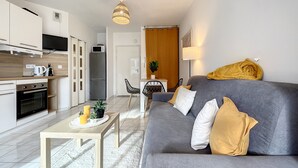 Un salon moderne avec un canapé gris, une table basse en bois et un coin cuisine doté de meubles blancs et d’appareils électroménagers.