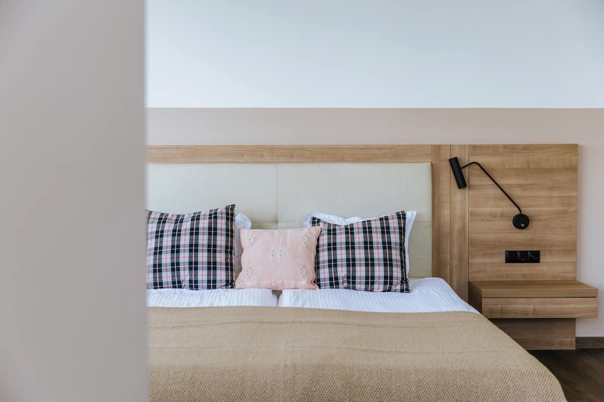 A neatly made bed with plaid and solid-colored pillows, a wooden headboard, and a bedside table with a lamp.