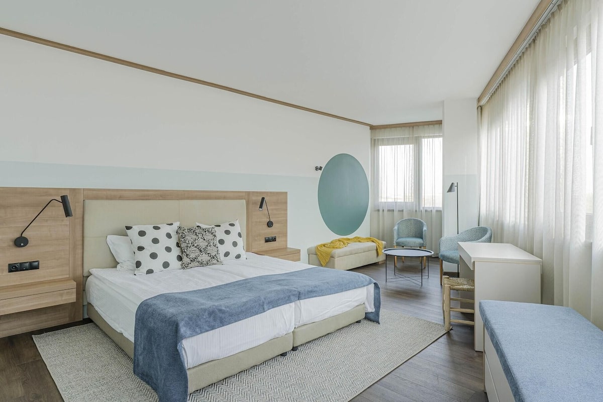 A modern hotel room with a large bed, a desk, a chair, and a sofa.