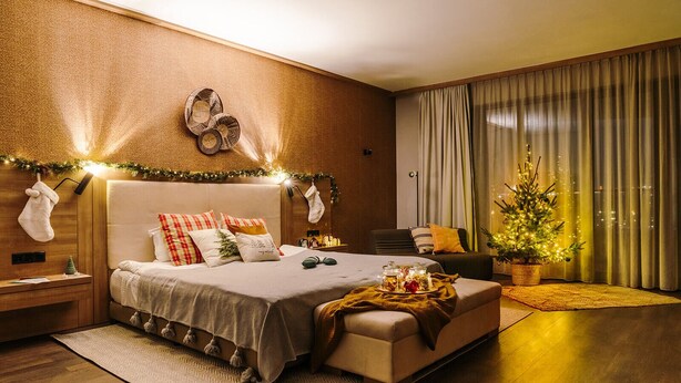 A modern bedroom with a large bed, a small side table, a nightstand, a chair, a Christmas tree, and a window with curtains.