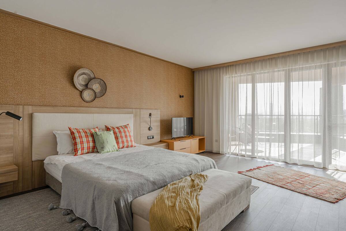 A modern hotel room with a large bed, a bedside table with a TV, a desk, and a large window with sheer curtains.