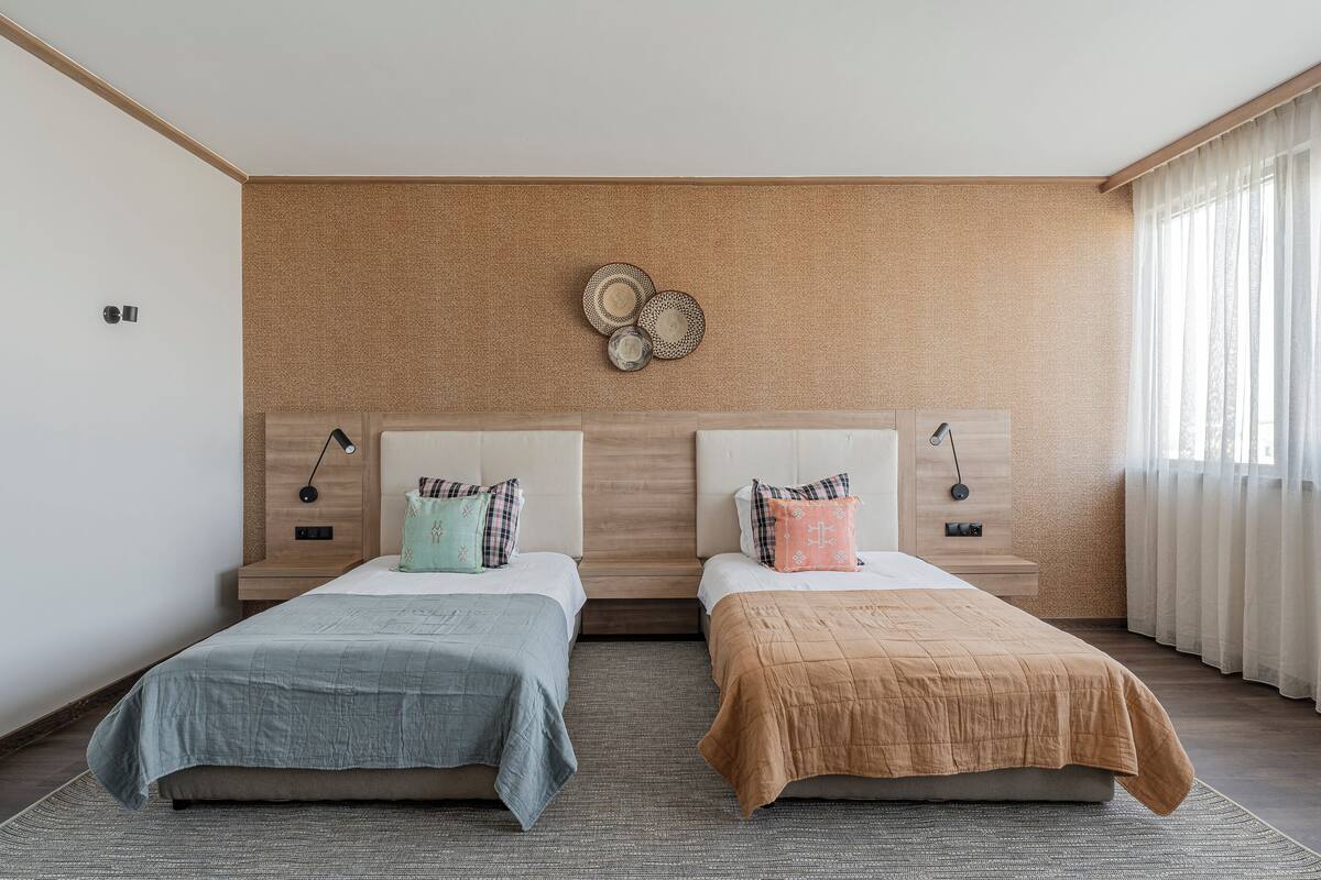 A modern hotel room with two beds, a wooden headboard, bedside lamps, and a large window with sheer curtains.