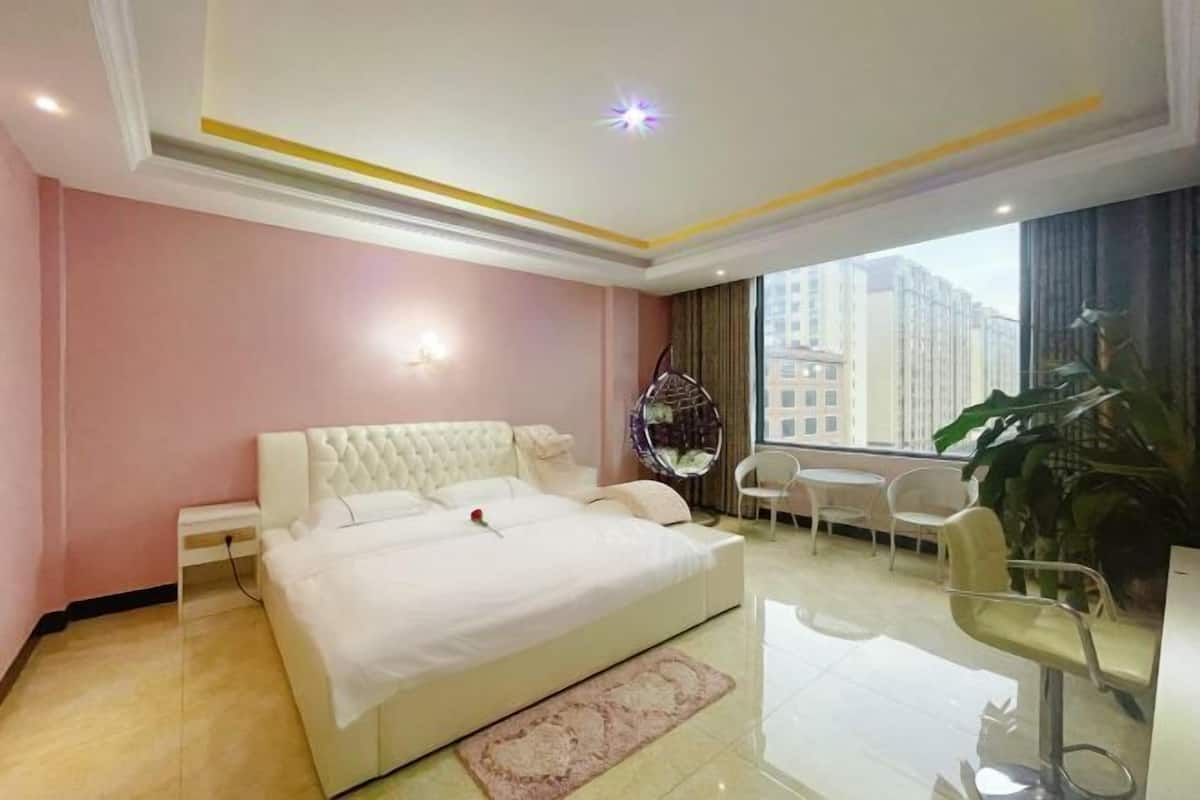 Moderni hotellihuone, jossa on suuri sänky, pieni pöytä, tuolit ja suuri ikkuna, josta avautuu näkymä kaupunkiin.
