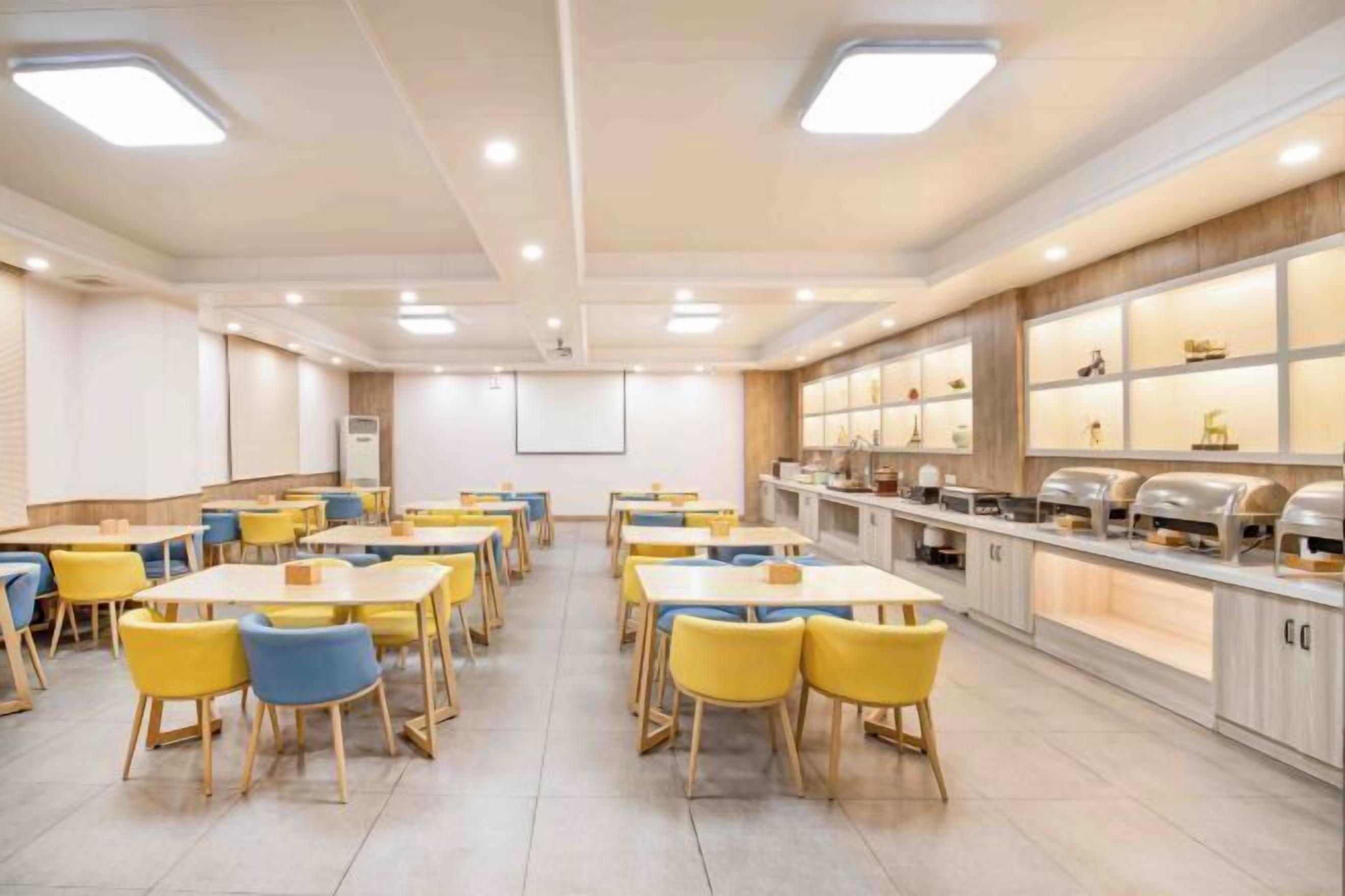 A spacious dining area with multiple tables and chairs, a buffet counter with various food items, and a projector screen on the wall.