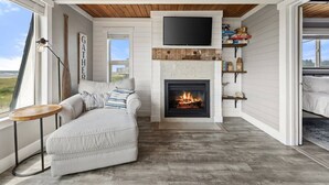 A cozy living room with a fireplace, a sofa, a TV mounted above the fireplace, and a view of the outdoors. - 100 Steps to the Beach, Amazing Views and Sunsets (Moclips)