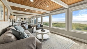 A spacious living room with a large window overlooking a beach, a cozy seating area, and a visible kitchen in the background. - 100 Steps to the Beach, Amazing Views and Sunsets (Moclips)