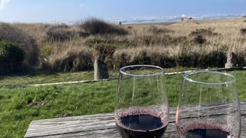 Deux verres à vin sur une table en bois, avec un champ verdoyant et une vue sur l’océan en arrière-plan.