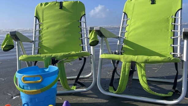 Dos sillas de playa verdes con estructura metálica y un cubo azul con juguetes de arena sobre una playa arenosa.