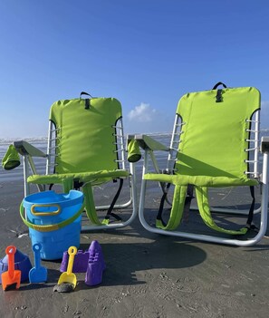 Two green beach chairs with metal frames and a blue bucket with sand toys on a sandy beach.