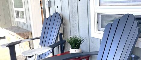 Een veranda met twee Adirondack-stoelen en een kleine tafel, een houten muur met een rustieke decoratie, en een raam met zonwering.