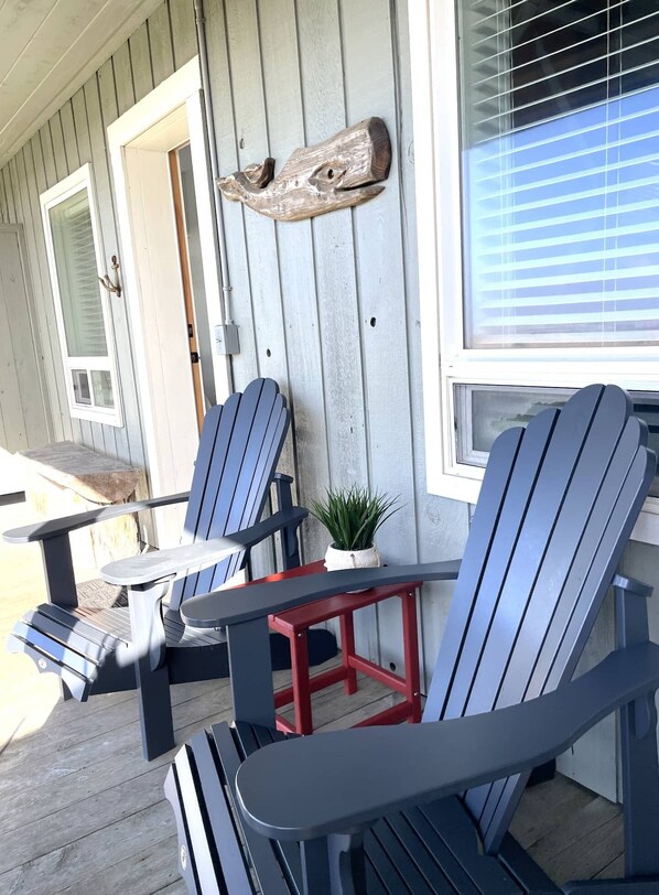 A porch with two Adirondack chairs and a small table, a wooden wall with a rustic decoration, and a window with blinds.