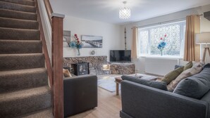 A living room with a staircase, a stone fireplace, a flat-screen TV, and a large window with curtains. - Sea Breeze Apt 2 - 2 Bed - Horton Beach (Swansea)