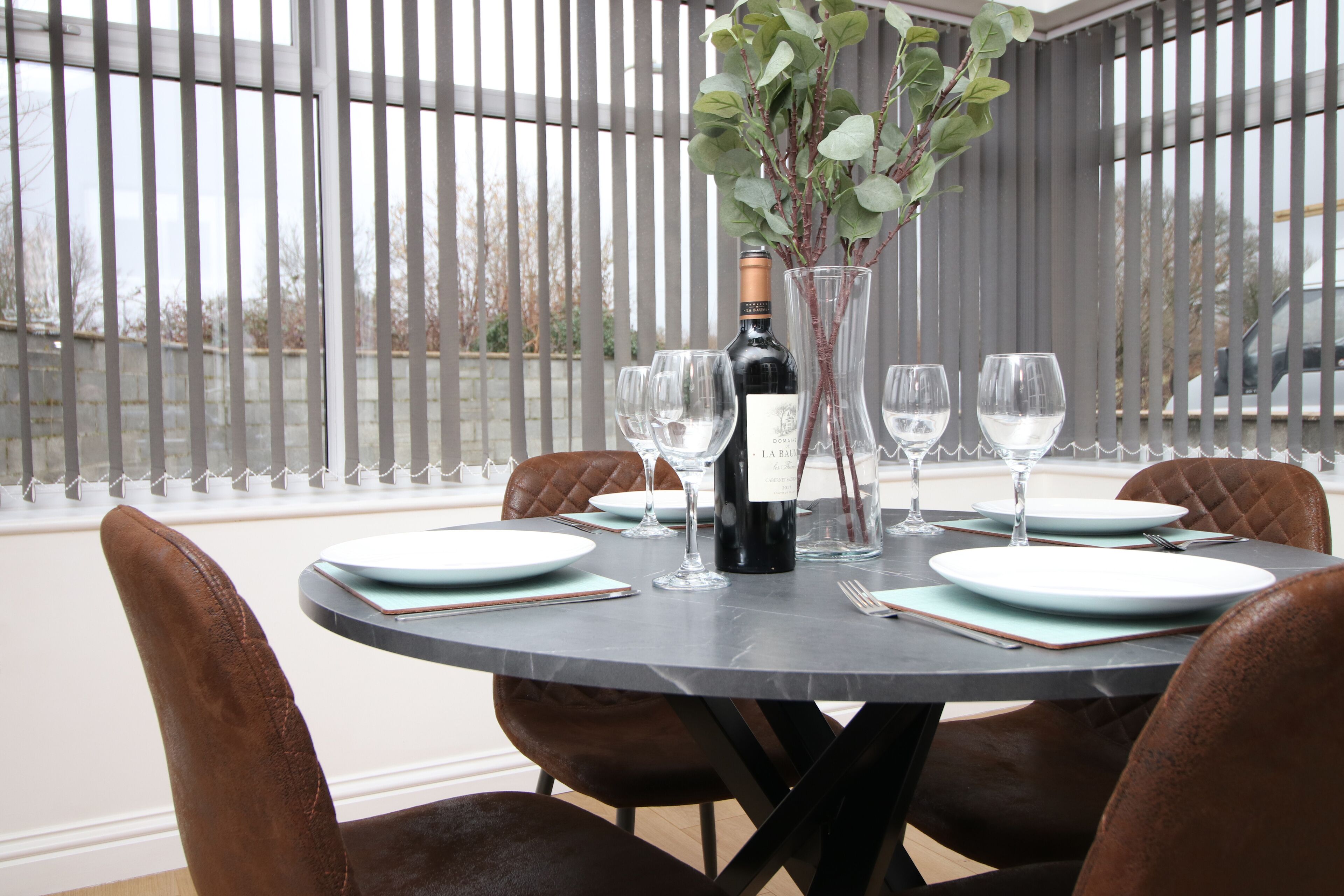 A round dining table with four brown leather chairs, a bottle of wine, and several wine glasses.