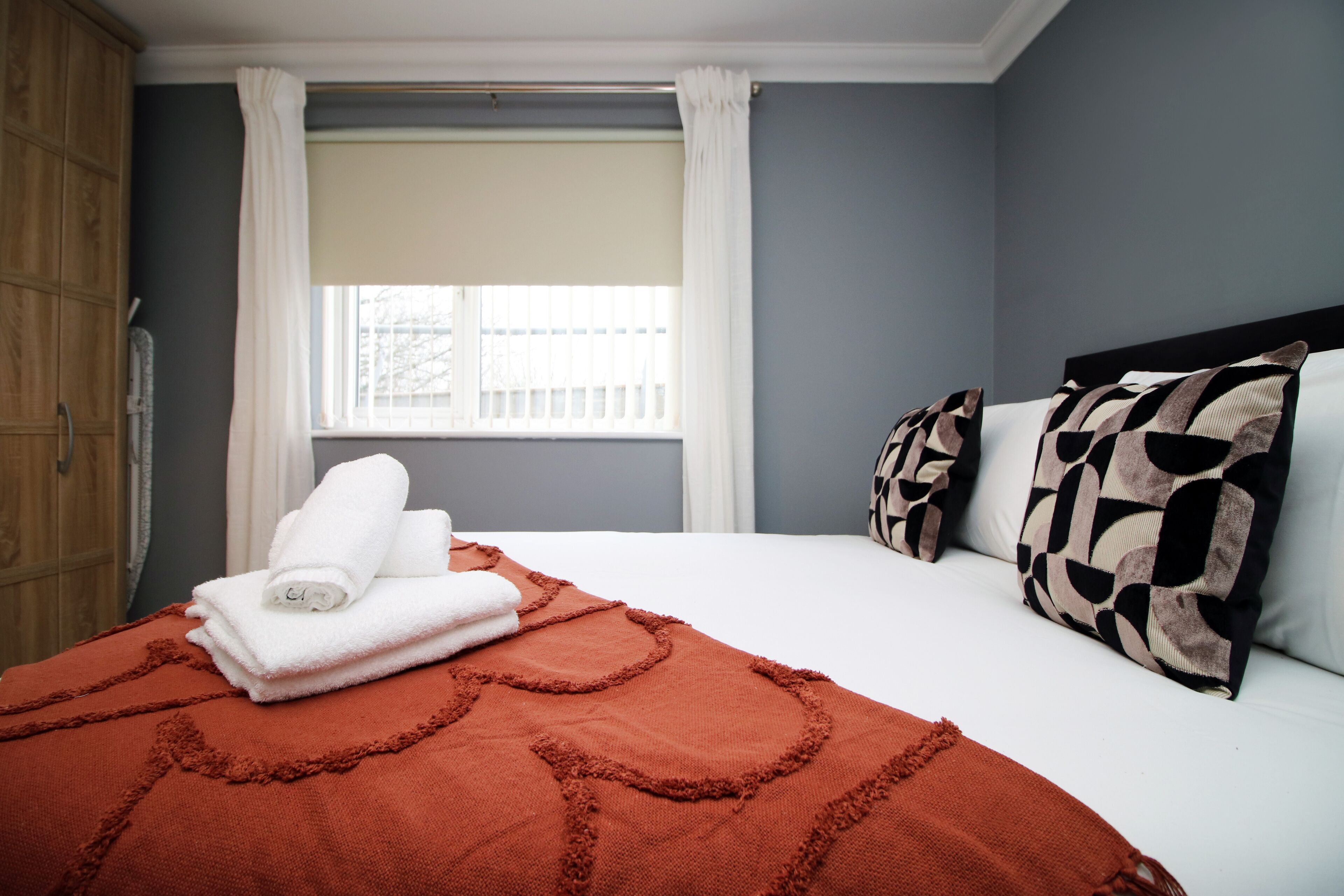 A neatly made bed with a red blanket and white towels, a window with white curtains, and patterned pillows.