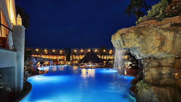 Vista nocturna de una piscina de hotel con cascada, rodeada de edificios iluminados y un paisaje rocoso.
