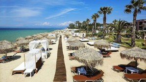 Un resort en la playa con filas de tumbonas y sombrillas de paja, un paseo marítimo de madera y palmeras.