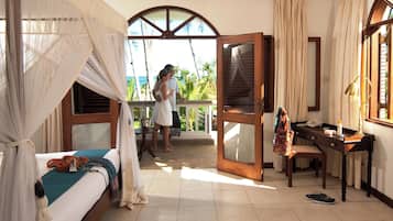 Quarto de hotel com cama, escrivaninha, cadeira, janela com vista para palmeiras e porta que dá para área externa.