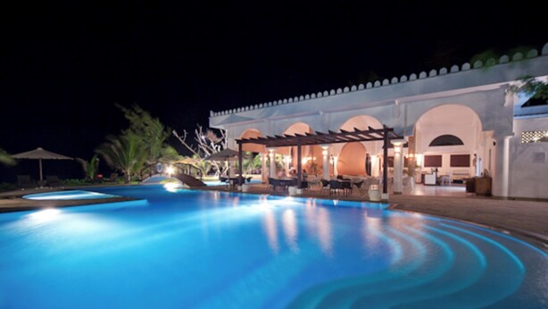 Ein Pool bei Nacht mit überdachter Sitzgruppe und einem Gebäude mit Bögen im Hintergrund.