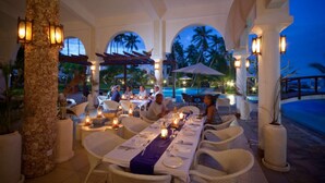 An outdoor dining area with tables set for a meal, surrounded by columns and potted plants. - Lantana Galu Beach (Diani Beach)